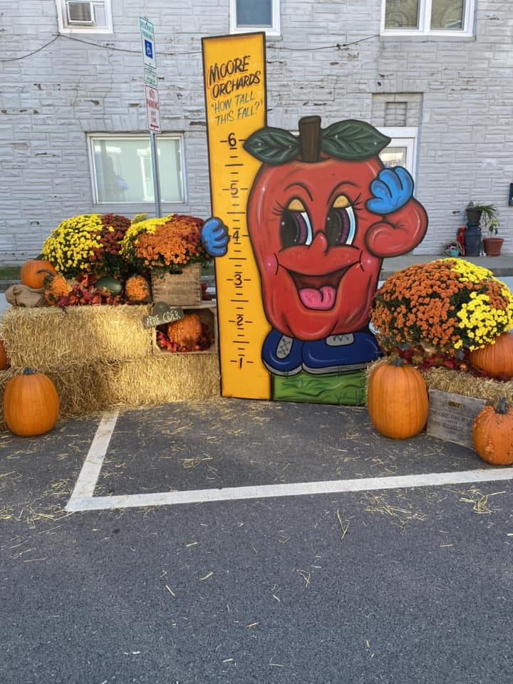 A smiling apple statue holding a measuring stick is surrounded by pumpkins and haystacks.