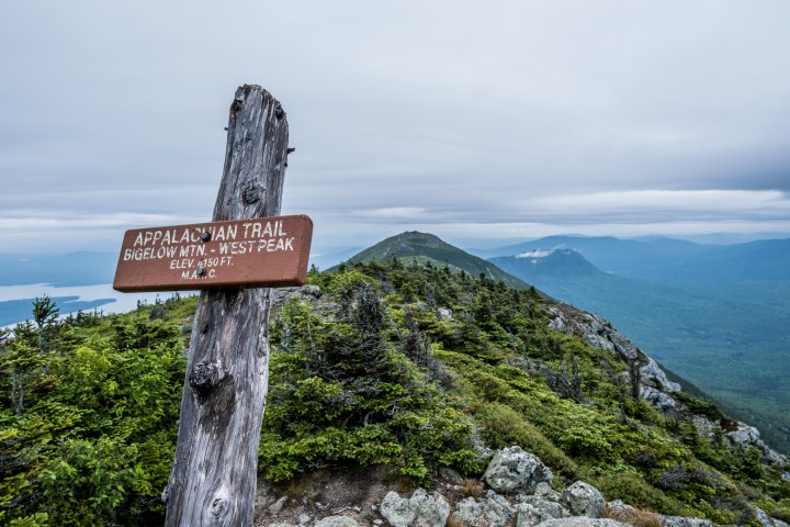 Standing above tree line on the Appalachian Trail in the mountains in Maine and looking at a trail sign.